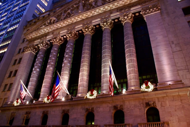 Fachada do prédio da Bolsa de Valores de Nova York iluminada à noite com uma grande bandeira dos Estados Unidos